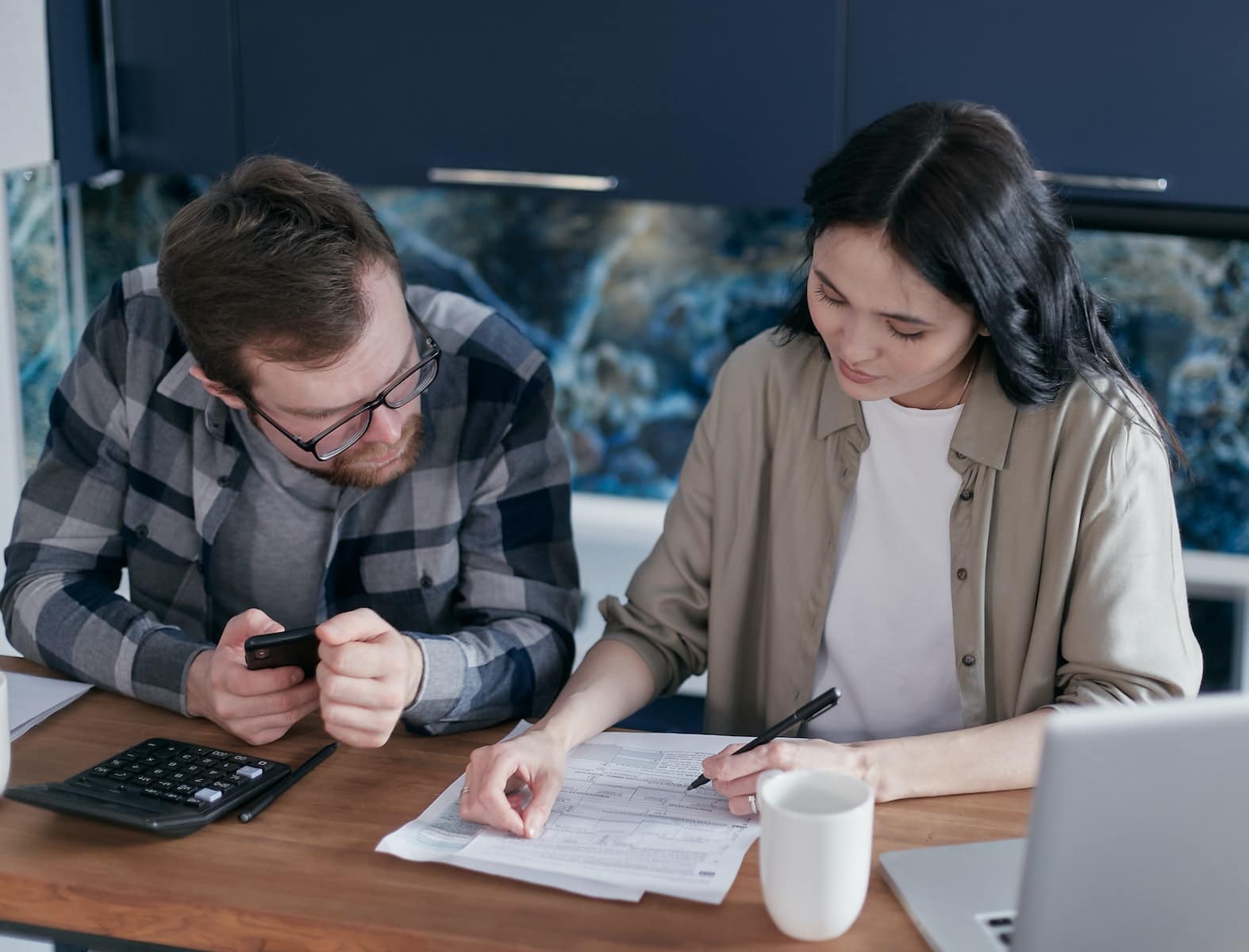 Man Holding a Cellphone Sitting Beside the Woman Signing Documents